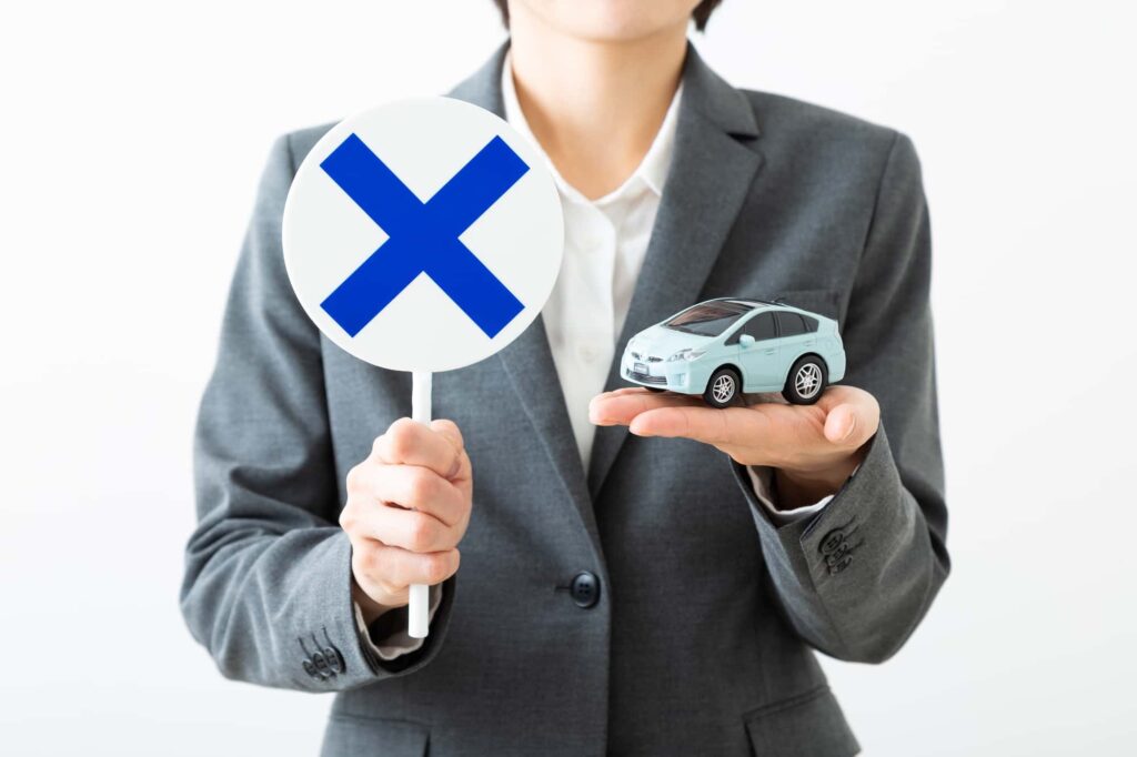 スーツの女性が×印の札とミニカーを持っている様子