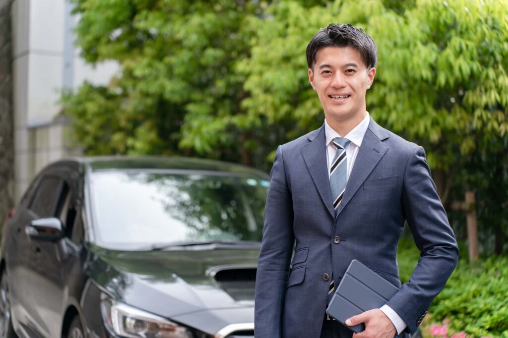 スーツ姿の男性が車の前で笑顔を見せている様子